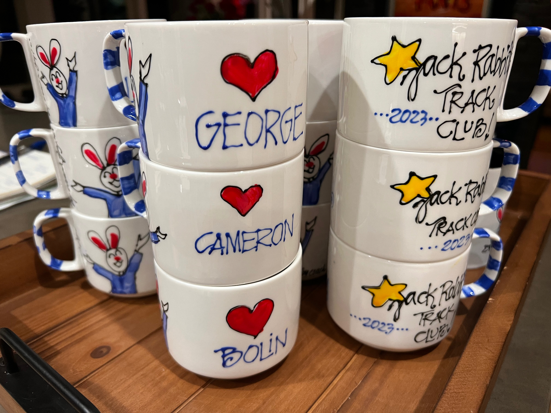 Stacks of hand painted mugs with kids' names like George, Cameron and Bolin beside Jack Rabbits Track Club 2023 mugs